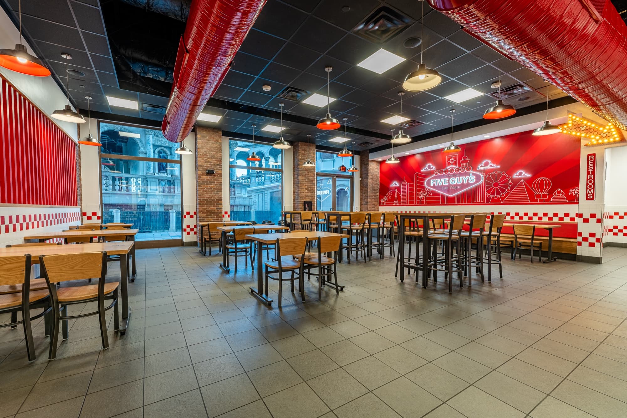Back dining room at Five Guys Grand Canal Shoppes, the buyout-friendly space: wood tables under red pendant lights, a Welcome to Fabulous Five Guys Las Vegas sign mural, checker-tile trim, and windows onto the Venetian streetscape