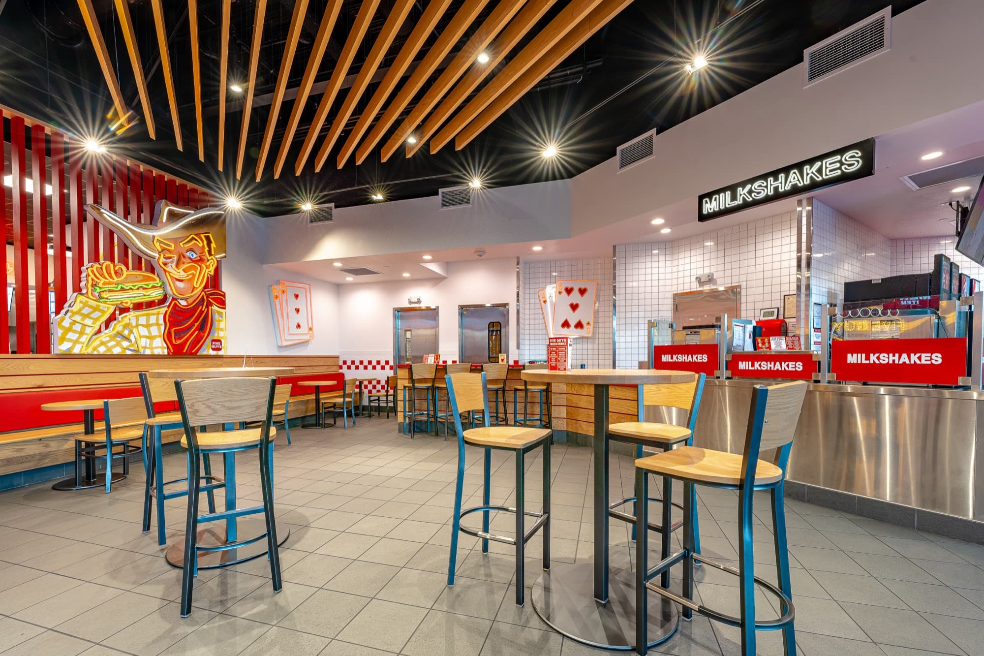 Shake bar at Five Guys Grand Canal Shoppes with a neon MILKSHAKES sign over a white-tile counter, high-top tables, and a neon cowboy mural holding a burger and fries