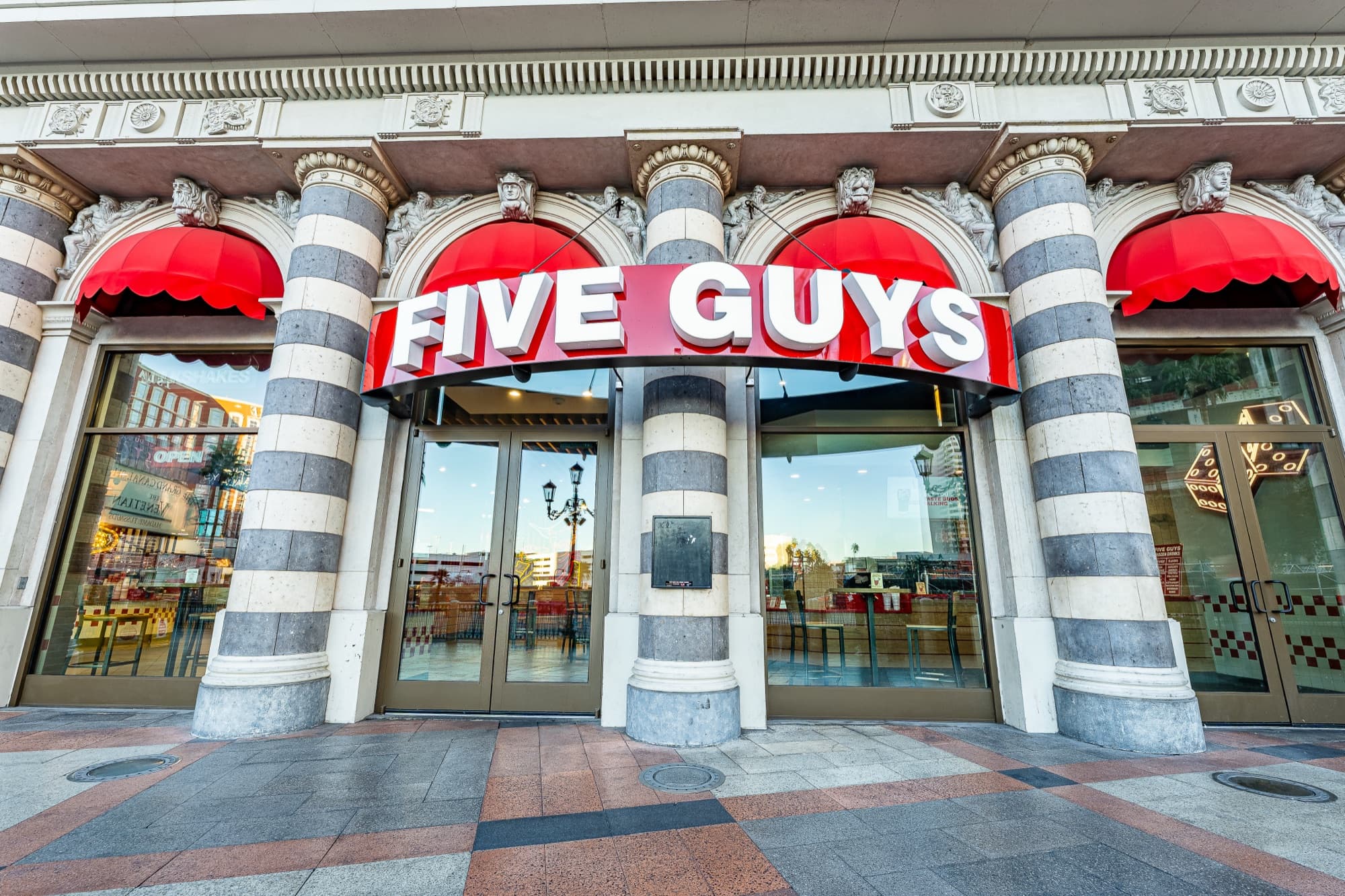 Five Guys storefront at Grand Canal Shoppes at The Venetian: red FIVE GUYS sign and scalloped red awnings between striped Venetian-style stone columns, glass doors open onto the promenade
