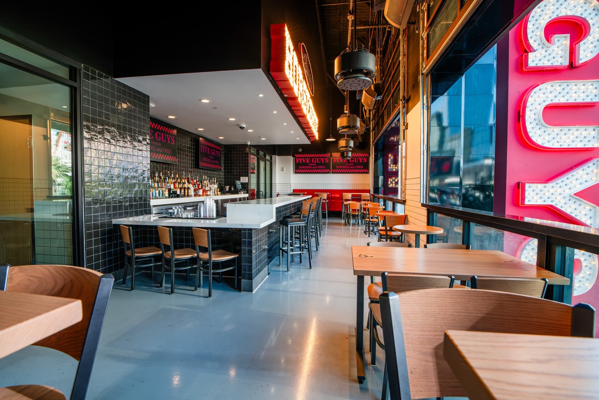 Wide view of the Five Guys Showcase bar room: white quartz bar, backlit GUYS marquee letters in the window wall, wood tables and red booths